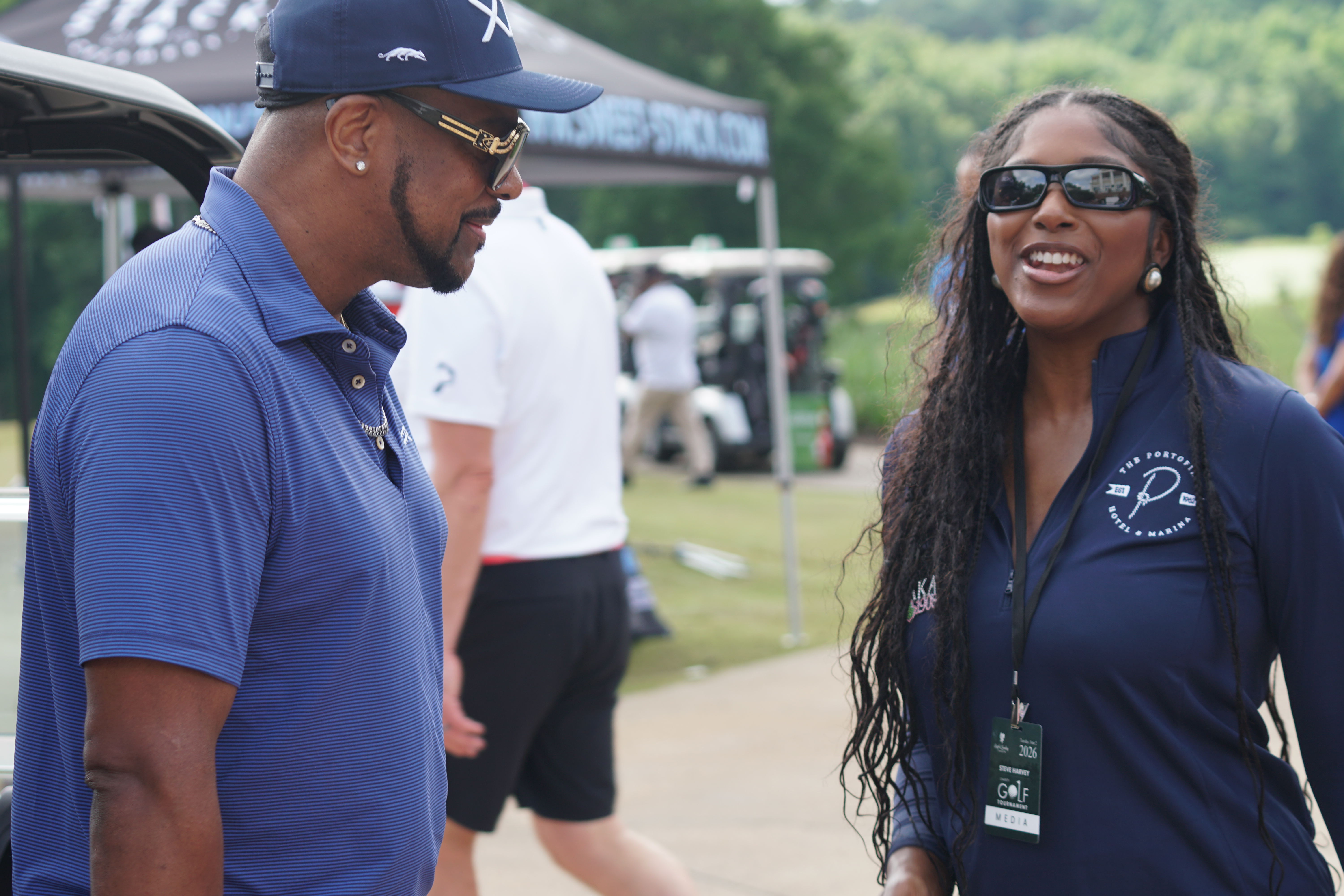Two people, a man and a woman, standing together and smiling at an outdoor event. The man is wearing a baseball cap and the woman is wearing sunglasses.