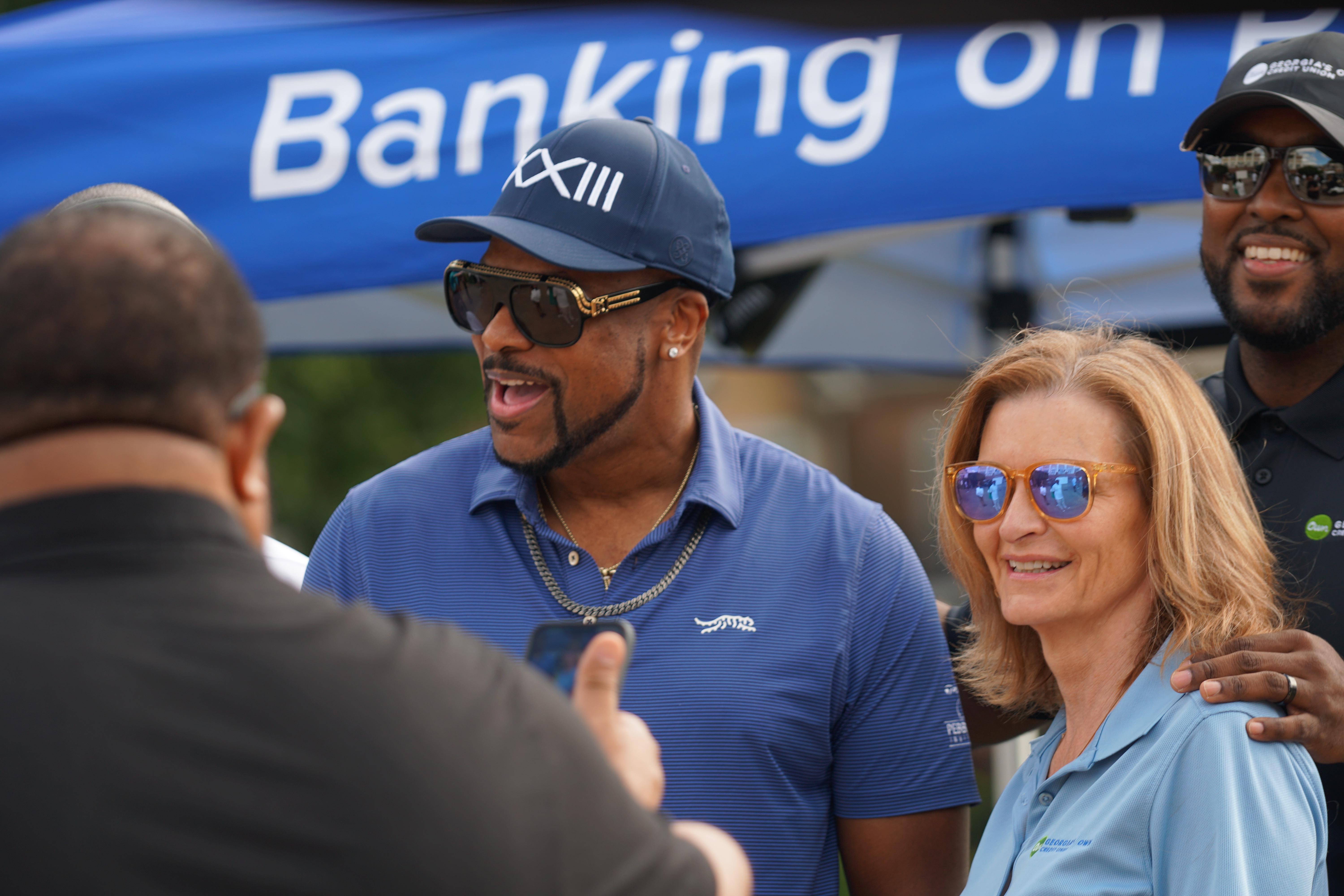 Two people in casual clothing standing in front of a "Banking on XIII" banner, one wearing a baseball cap and sunglasses.
