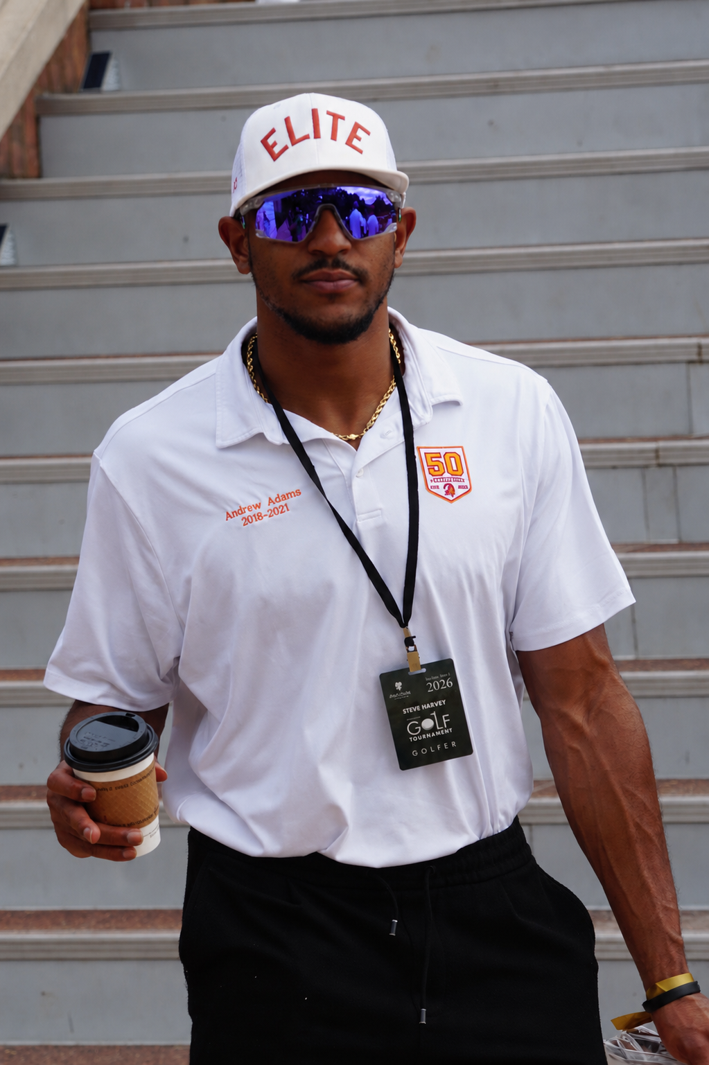 A man wearing a white shirt with "ELITE" printed on it, sunglasses, and a lanyard standing on stairs.
