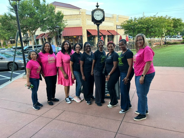 A group of women wearing pink and black shirts standing together in front of a commercial building with a clock tower.