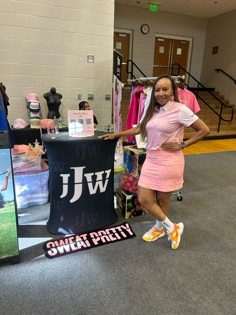 A woman in a pink dress standing next to a display table with "JHW" and "Sweat Pretty" branding.