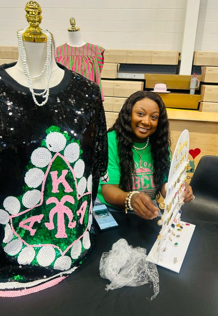 A smiling woman wearing a green shirt and holding a sign with the letters "Kappa Alpha Psi" in front of a display of sequined clothing.