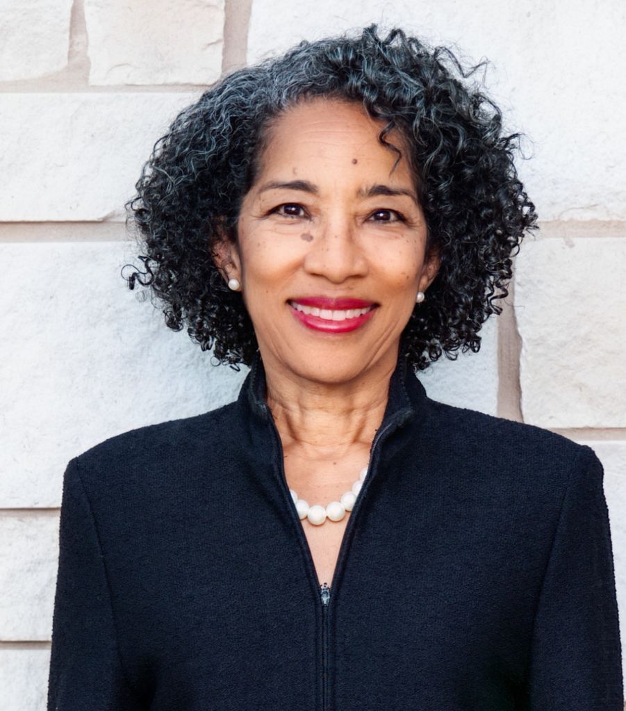 A smiling older woman with curly black hair wearing a black jacket and pearl necklace standing in front of a white wall.