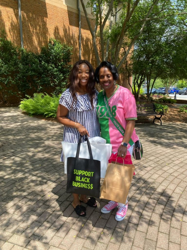 Two smiling women, one wearing a pink shirt with "918" and the other in a striped dress, standing in front of a brick building and holding a "Support Black Business" tote bag.