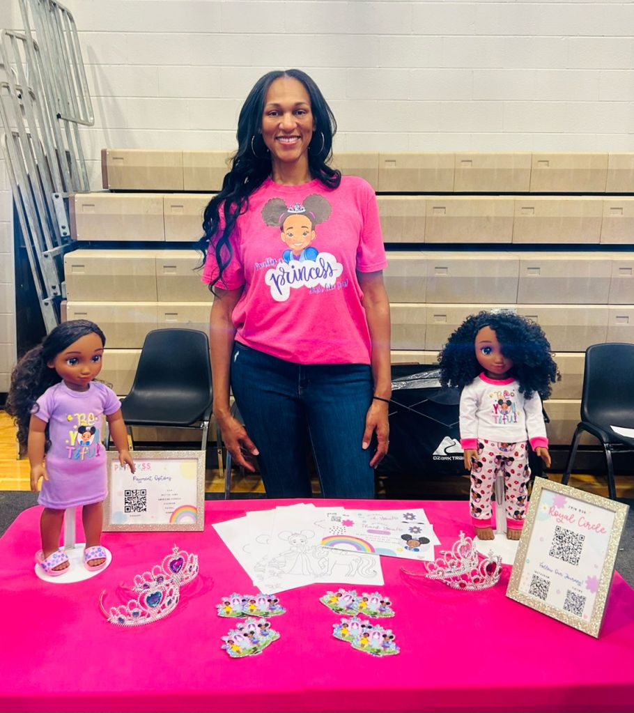 A smiling woman wearing a pink "Princess" t-shirt stands with two Black dolls on a table displaying princess-themed items.