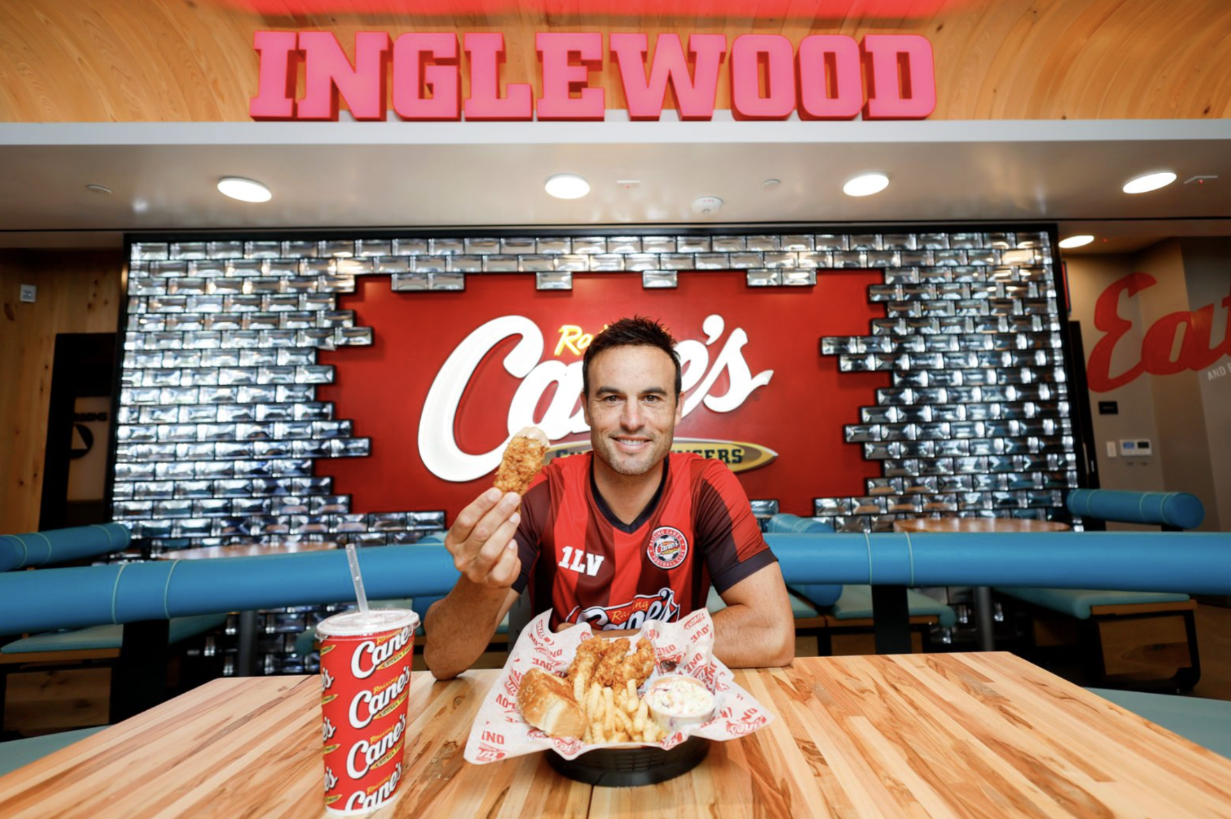 A man in a red sports jersey holding a large order of food from Carl's Jr. restaurant, sitting in front of a Carl's Jr. logo and Inglewood sign.