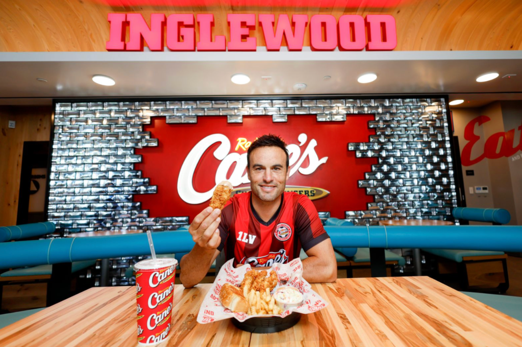 A man in a red sports jersey holding a large order of food from Carl's Jr. restaurant, sitting in front of a Carl's Jr. logo and Inglewood sign.