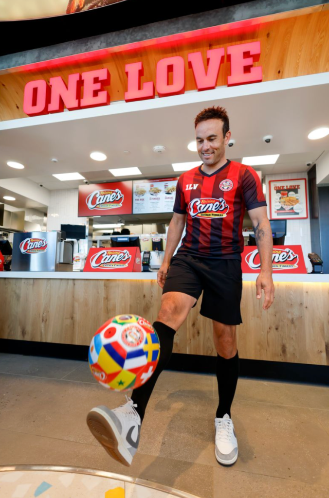A smiling man in a Cane's restaurant uniform stands in front of a "One Love" sign, holding a colorful soccer ball.
