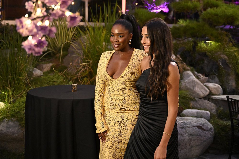 Two women in formal attire posing together in a garden setting with colorful flowers and lighting.