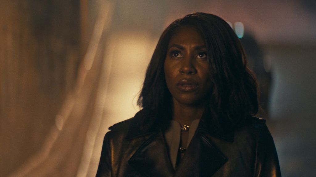 A woman with long dark hair wearing a black coat stands in a dimly lit setting, her expression serious.