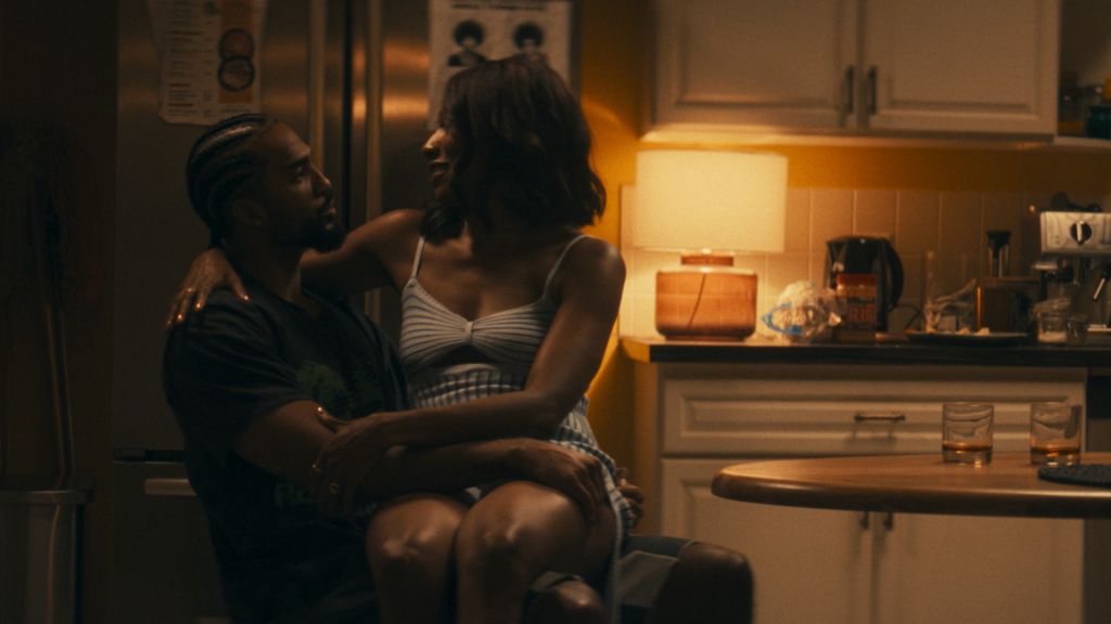 Two people embracing intimately in a dimly lit kitchen, with a coffee maker and other appliances visible in the background.