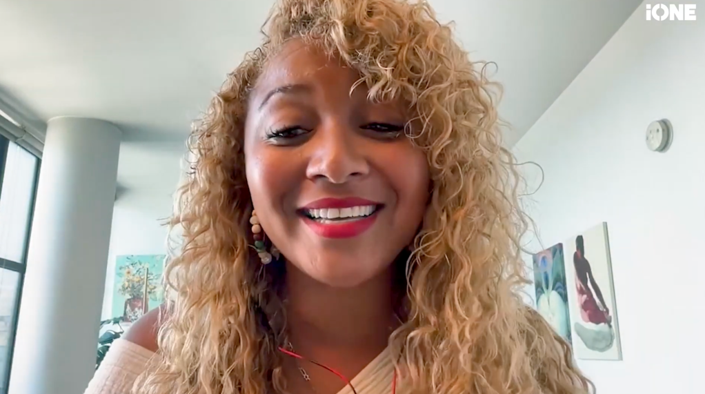 A smiling woman with curly blonde hair wearing a pink top and earrings.