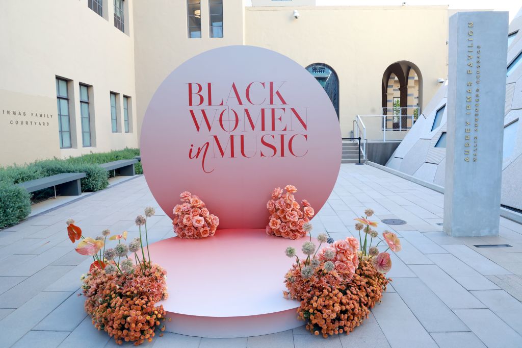 A large circular display with the text "BLACK WOMEN in MUSIC" surrounded by pink and orange floral arrangements, set against a modern architectural backdrop.
