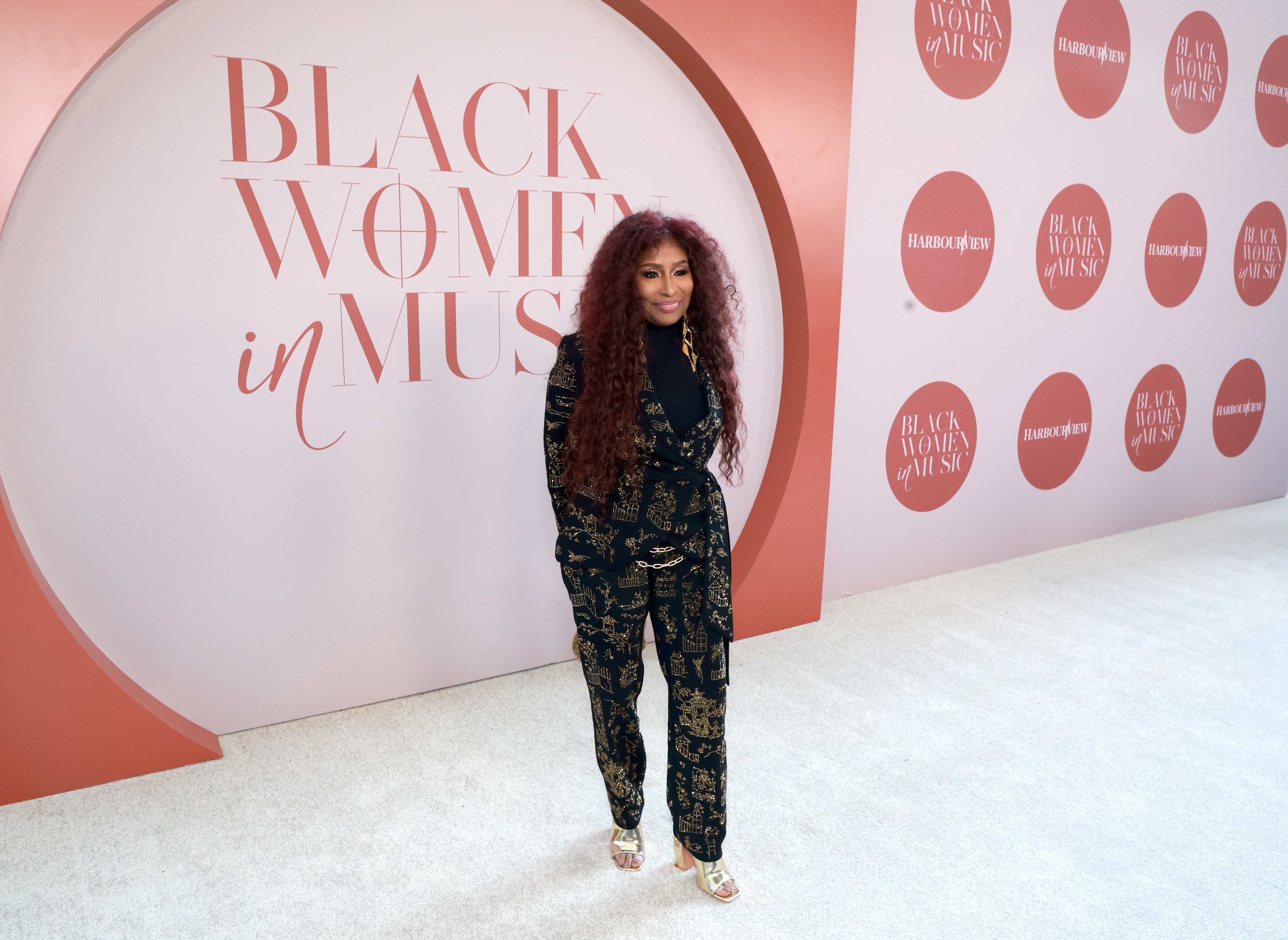 A woman with curly red hair wearing a black and gold patterned jumpsuit stands in front of a pink backdrop with the text "Black Women in Music".
