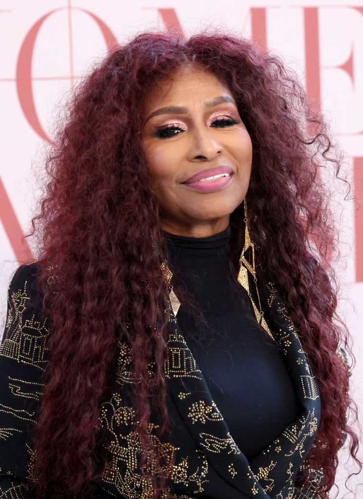A woman with curly burgundy hair wearing a black and gold patterned jacket smiling at the camera.