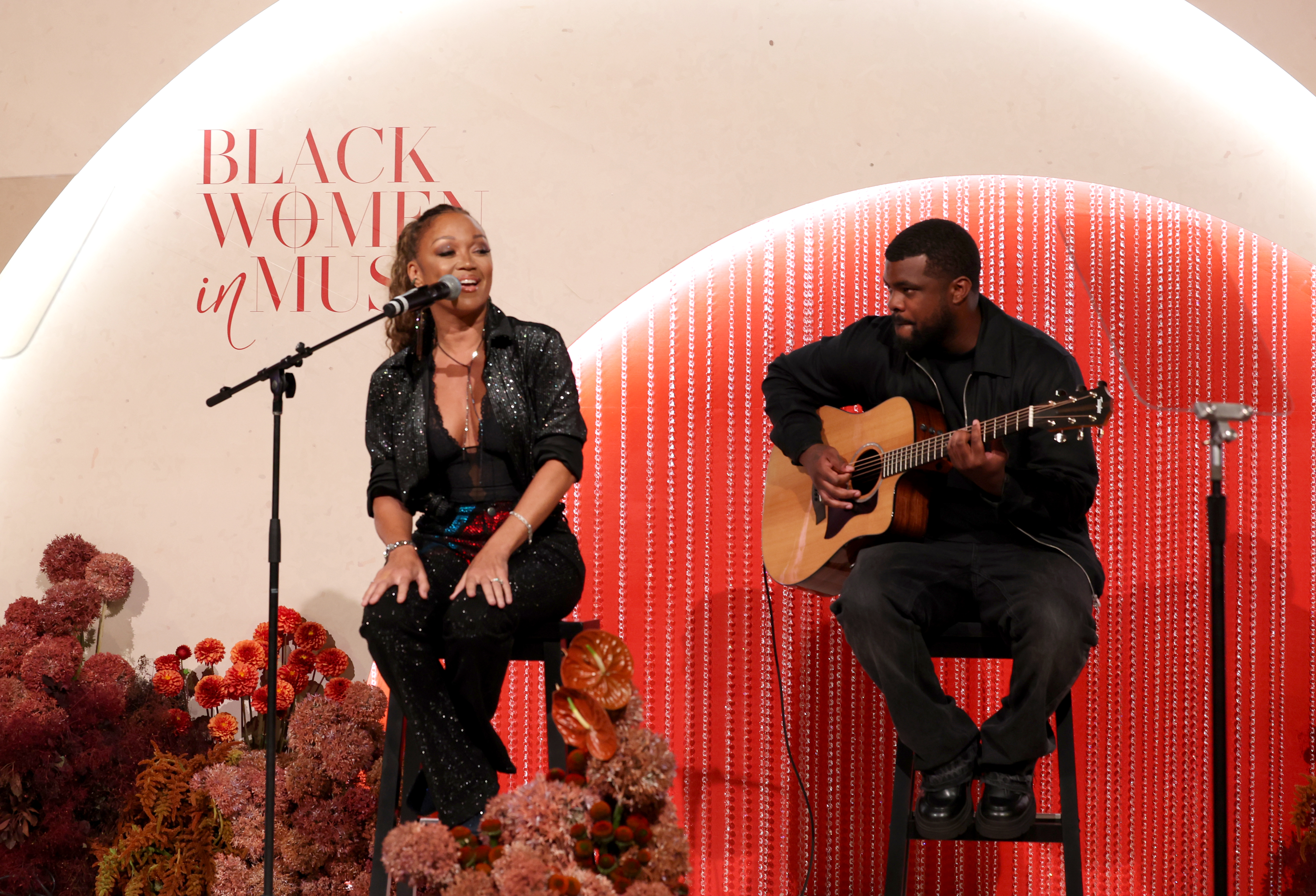 Two people performing on stage, with a woman singing into a microphone and a man playing an acoustic guitar. The stage is decorated with red and pink flowers and a backdrop that says "Black Women in Music".