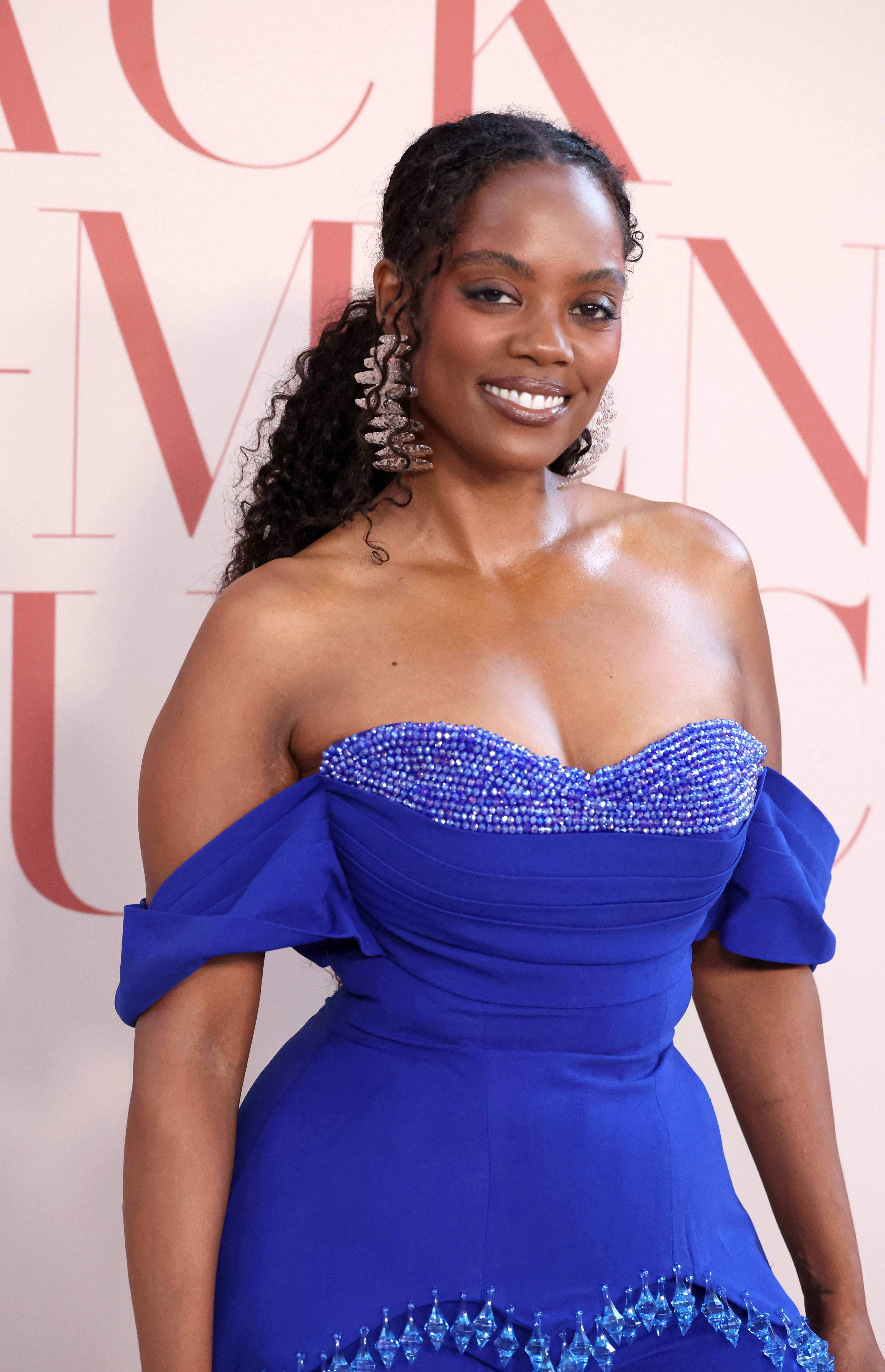 A smiling woman with curly dark hair wearing a blue off-the-shoulder dress with beaded embellishments, standing in front of a pink and white background.