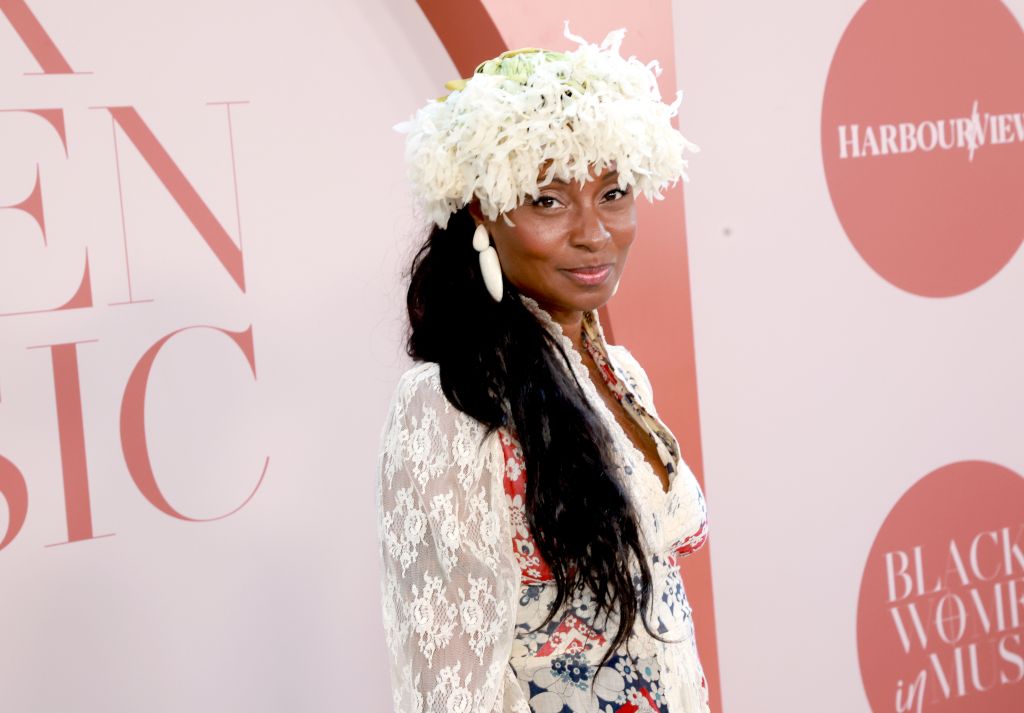 A woman wearing a white lace dress and a feathered headdress stands in front of a pink backdrop with "Harbour View" text.