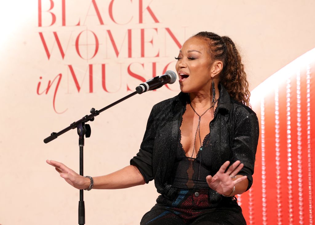 A woman singing passionately into a microphone on stage, with the text "BLACK WOMEN IN MUSIC" displayed behind her.