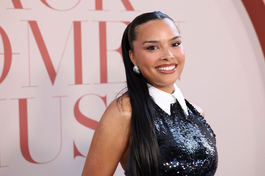 A smiling woman with long dark hair wearing a sequined black dress and white collar stands in front of a pink backdrop.