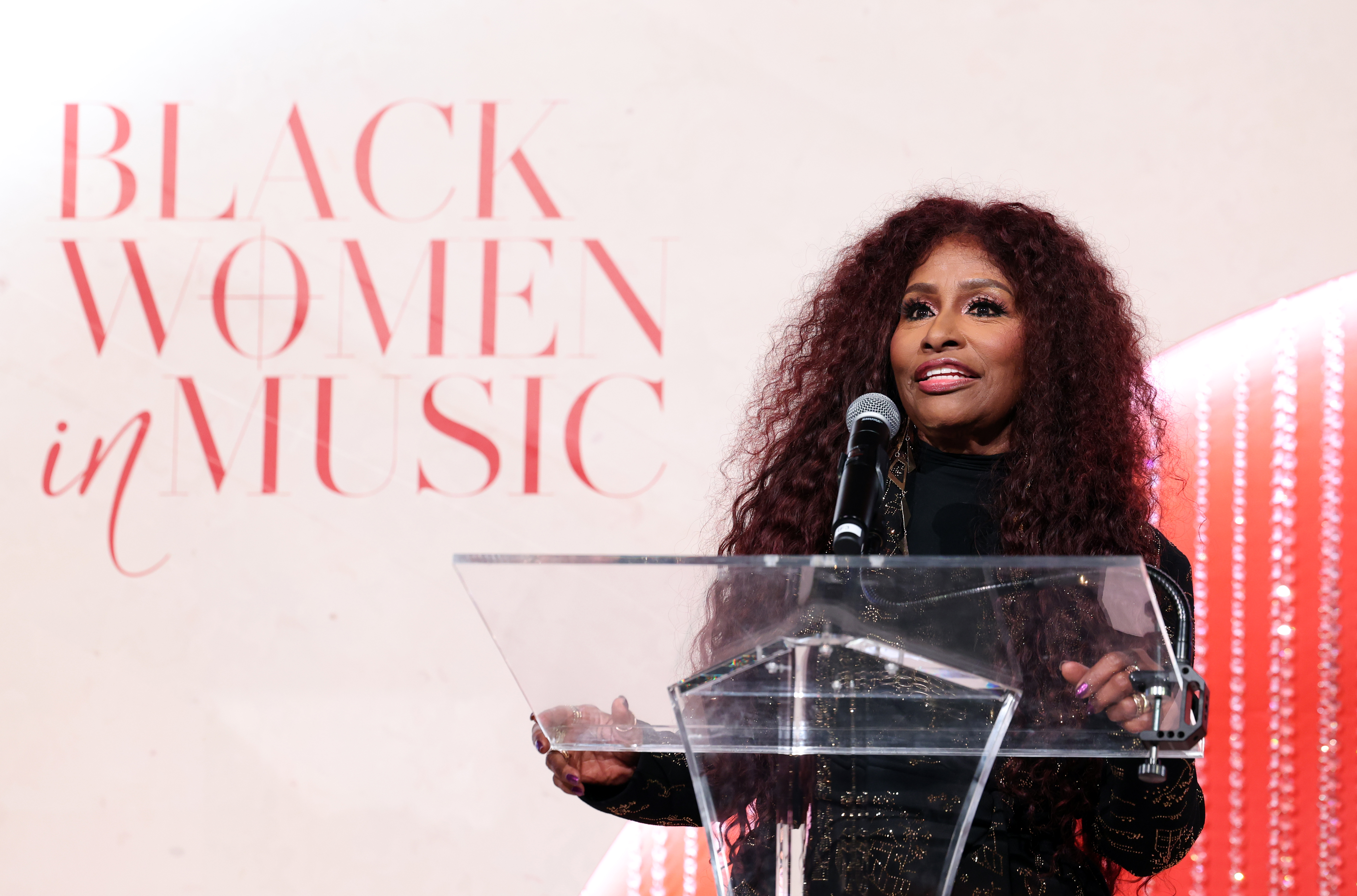 A woman with curly red hair speaking into a microphone at a podium with the text "Black Women in Music" displayed behind her.