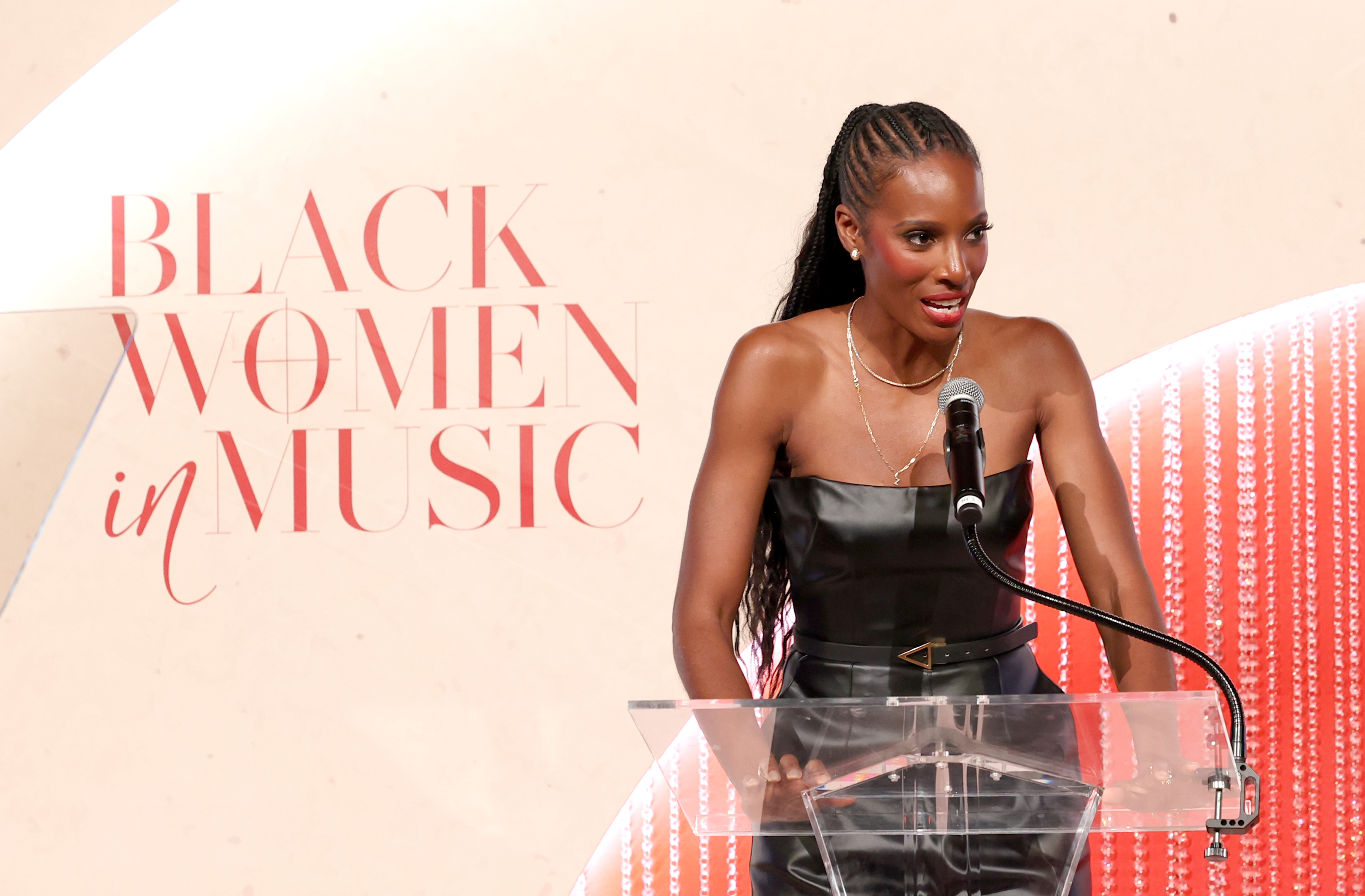 A woman speaking at a podium on stage, with a backdrop displaying the text "Black Women in Music".