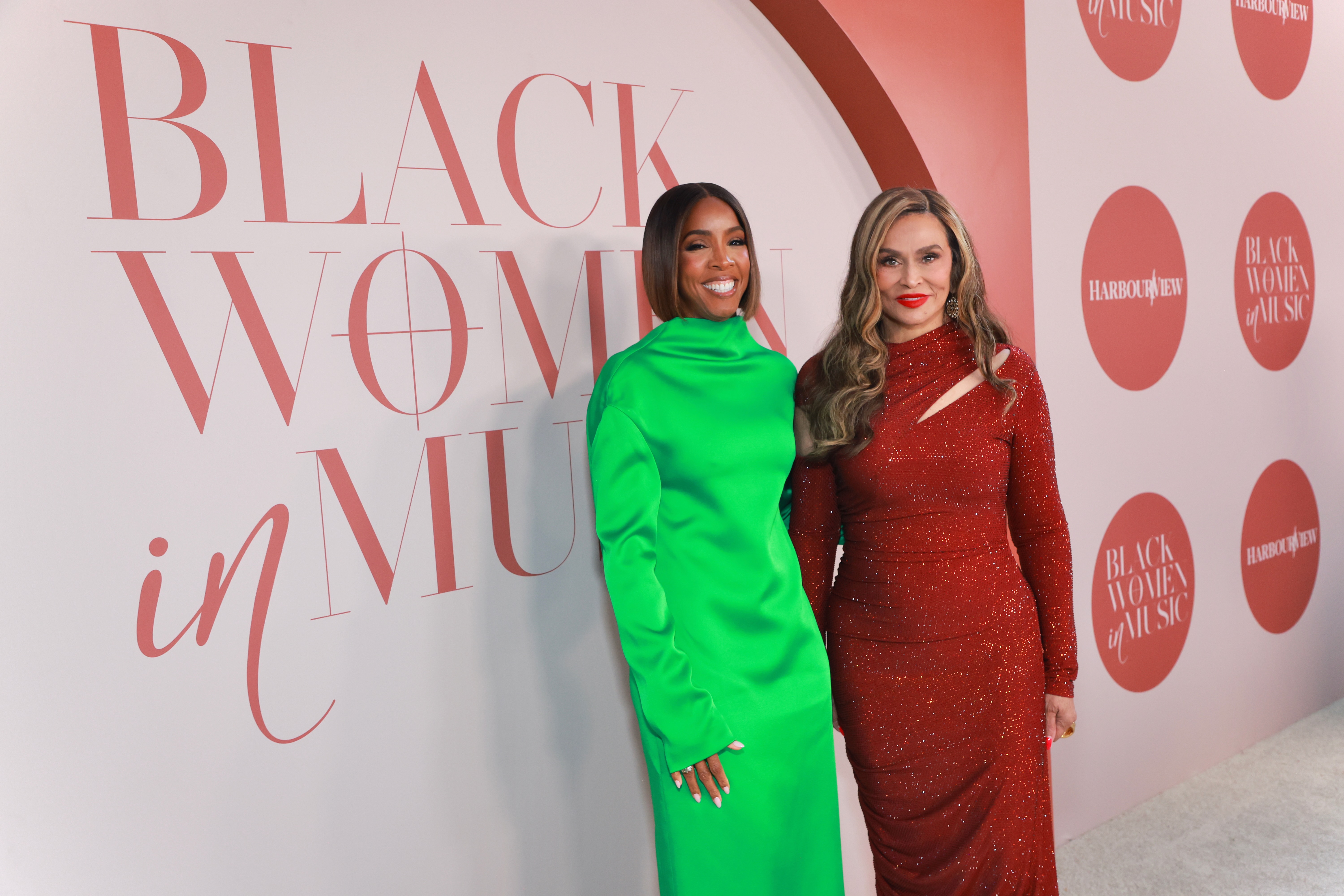 Two women posing in front of a "Black Women in Music" backdrop, one wearing a bright green dress and the other a sparkling red dress.