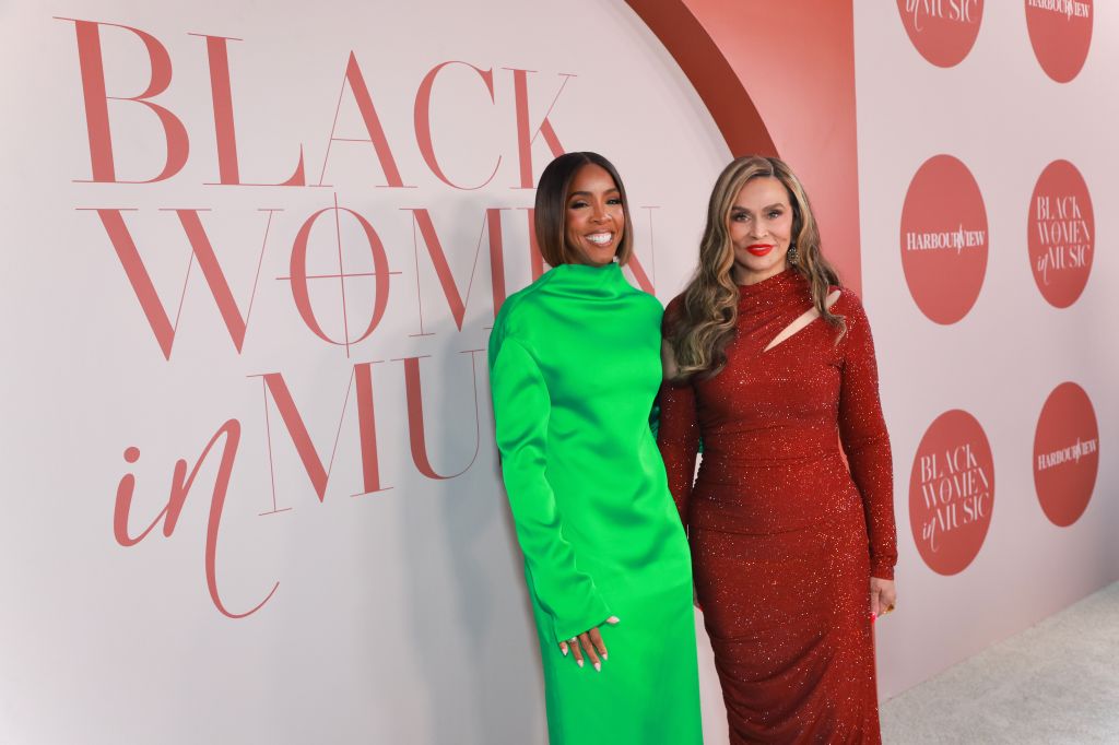 Two women posing in front of a "Black Women in Music" backdrop, one wearing a bright green dress and the other a sparkling red dress.