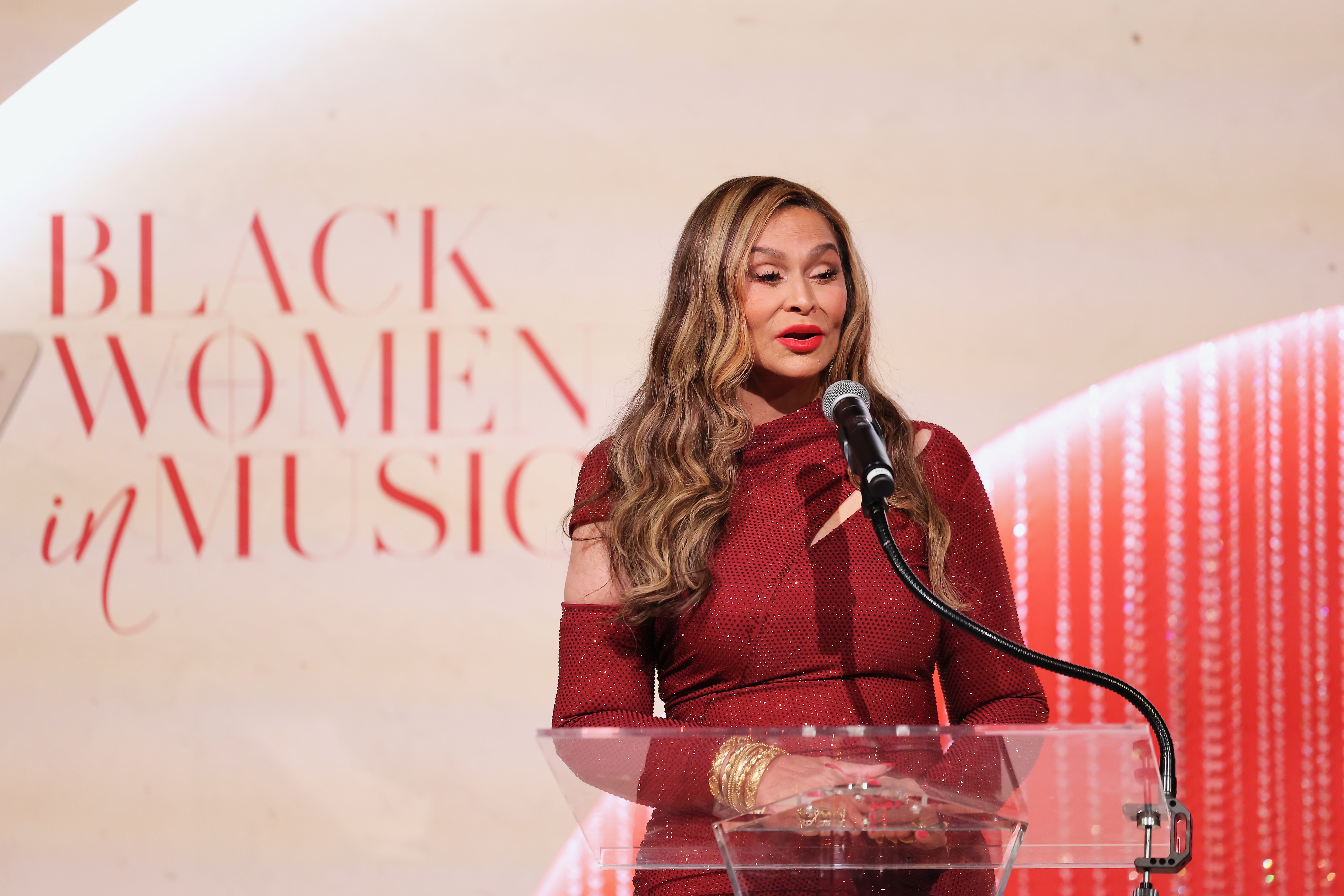 A woman in a red sequined dress speaking into a microphone at a podium with the text "Black Women in Music" displayed behind her.