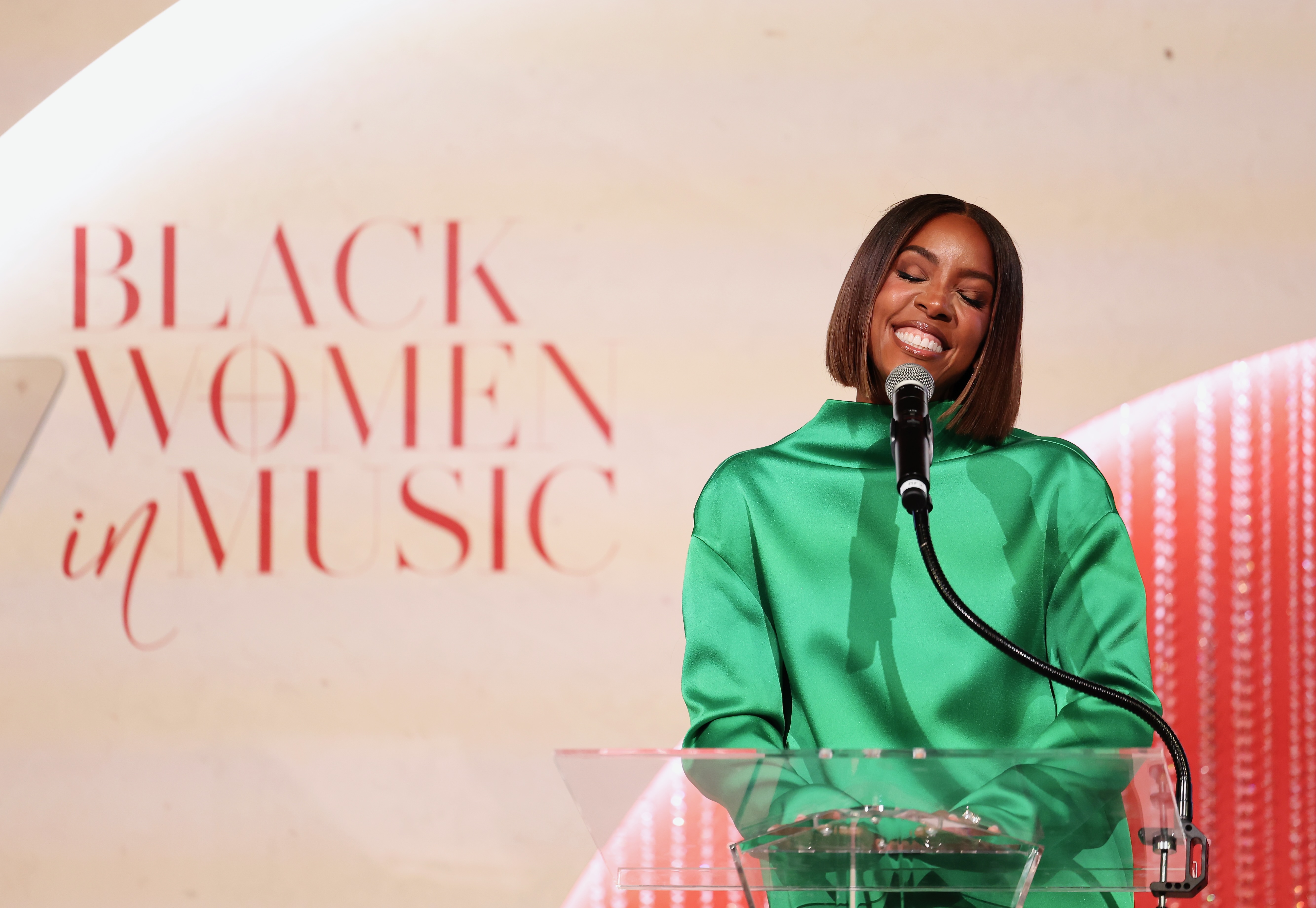 A woman in a green dress speaking into a microphone on a stage, with a "Black Women in Music" banner visible behind her.