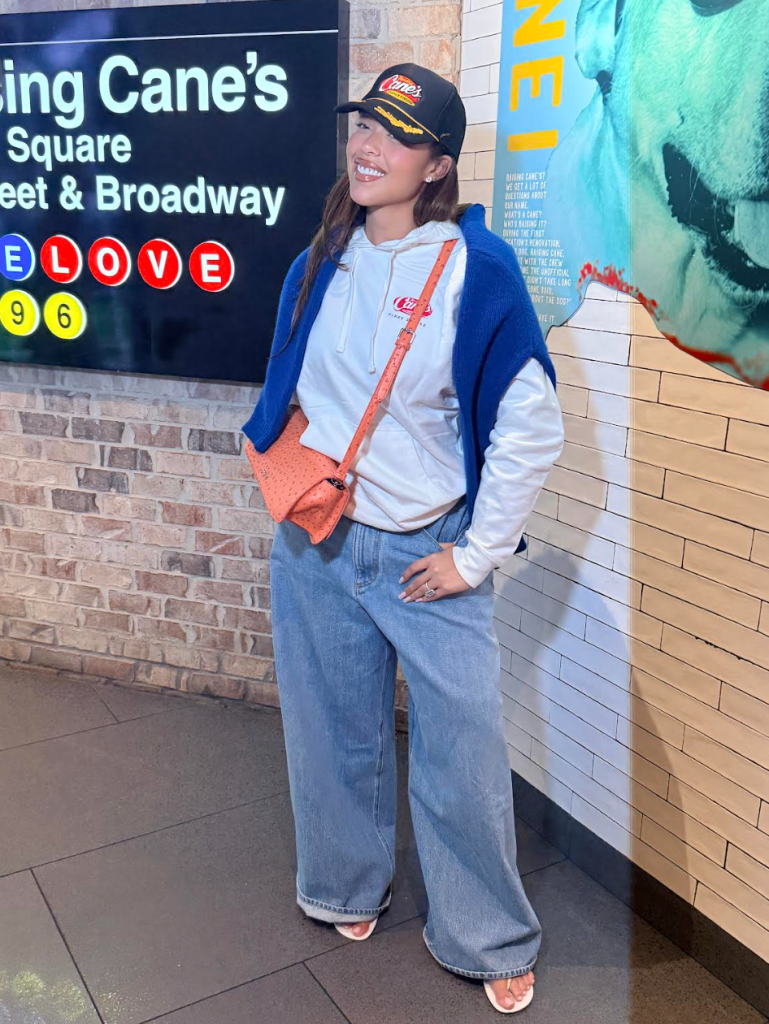 A smiling young woman wearing a Raising Cane's cap and hoodie stands in front of a Raising Cane's Square Street & Broadway sign.