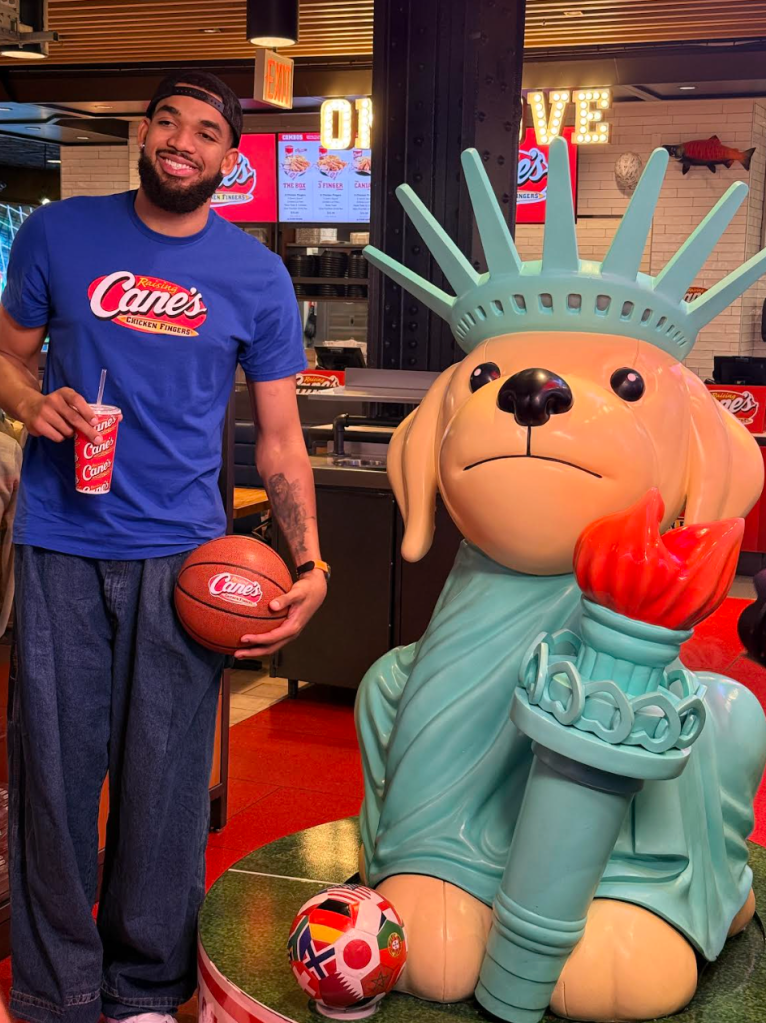 A smiling man in a Cane's Chicken Fingers t-shirt holding a basketball in front of a large Statue of Liberty figure.