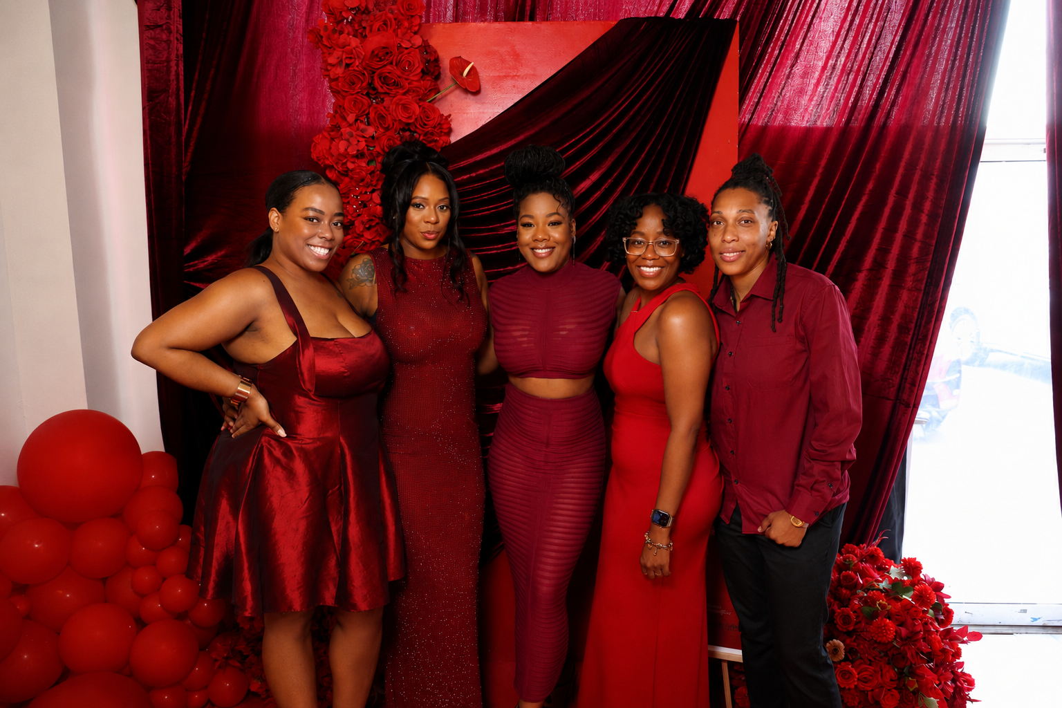 A group of five young women dressed in red formal attire posing in front of a red and black backdrop with red floral arrangements.