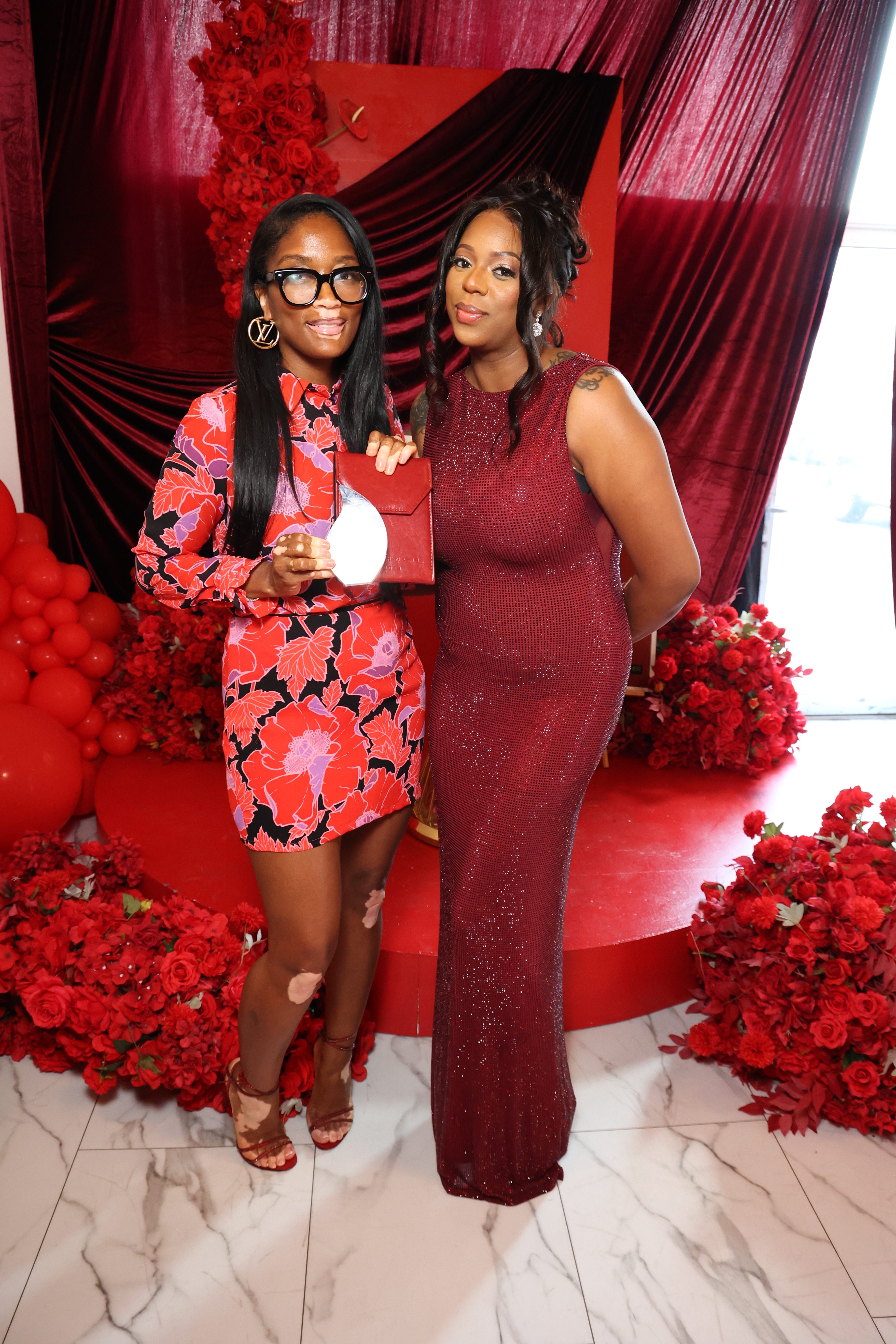 Two women in formal red dresses posing in front of a red floral backdrop, one wearing glasses and holding a red purse.