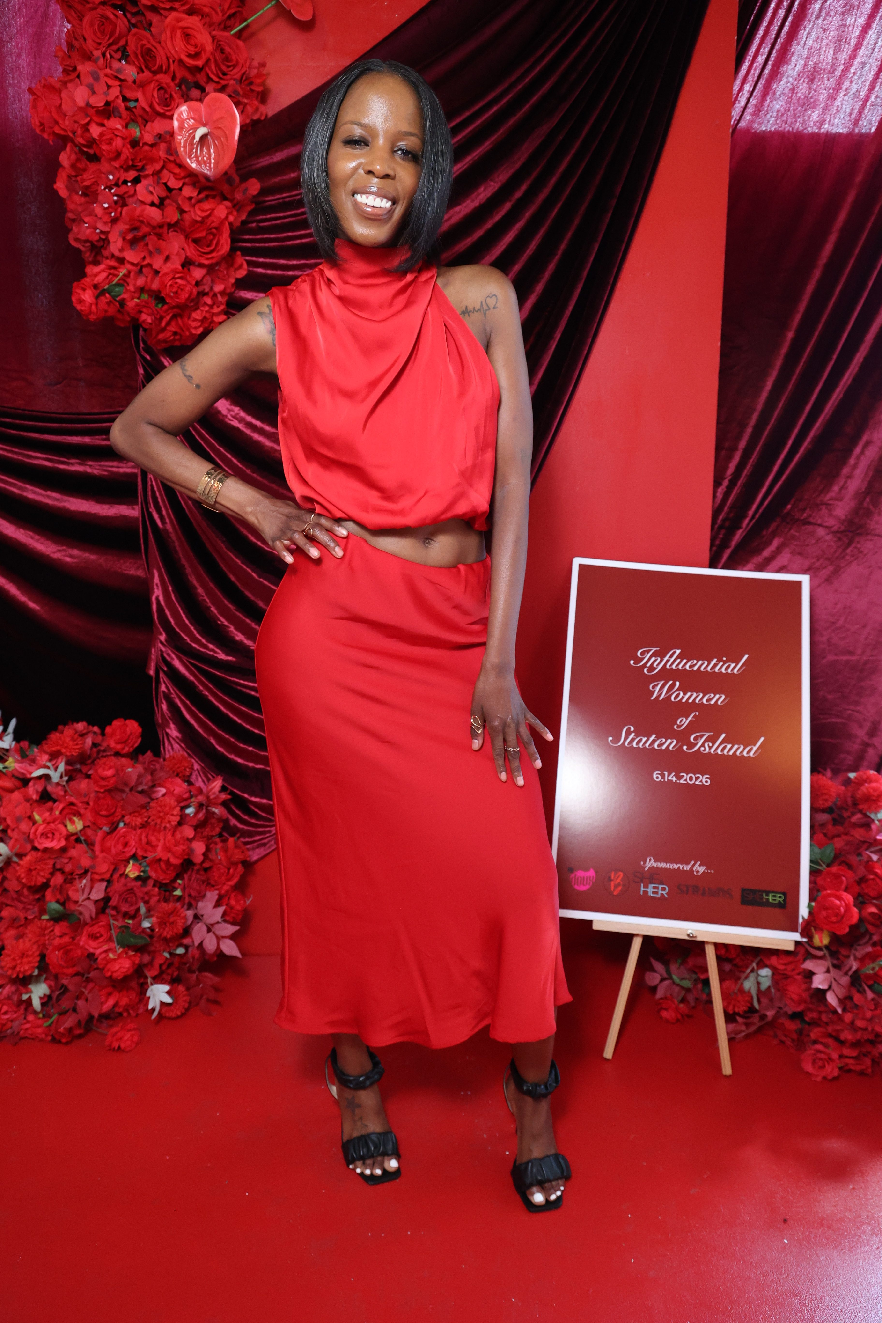 A smiling woman in a red dress stands in front of a red floral backdrop, holding a sign that reads "Influential Women of Staten Island 6.14.2026".