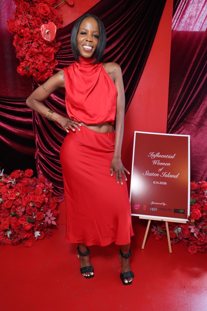 A smiling woman in a red dress stands in front of a red floral backdrop, holding a sign that reads "Influential Women of Staten Island 6.14.2026".