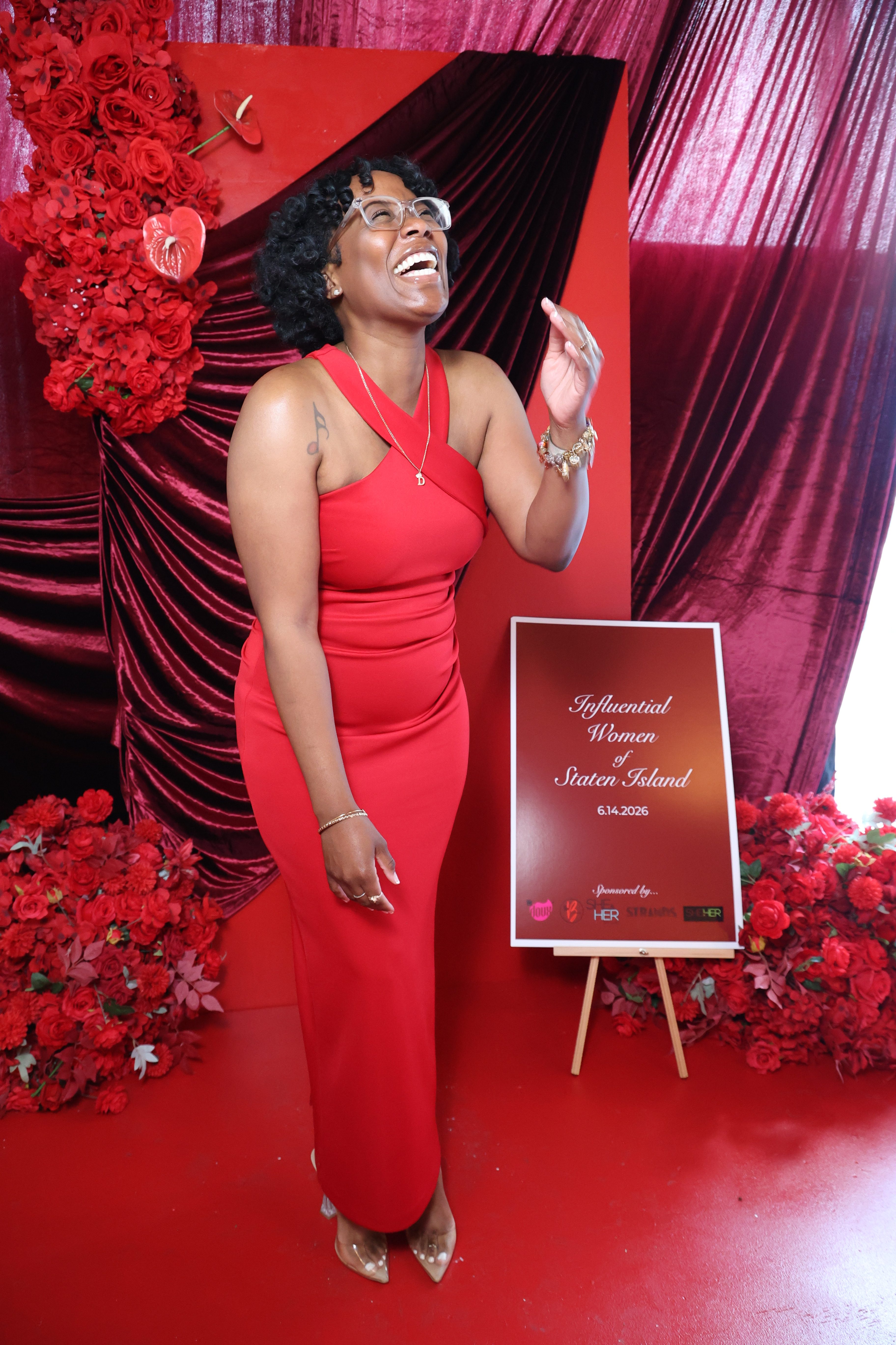 A smiling woman in a red dress stands in front of a red and black backdrop, holding a sign that reads "Influential Women of Staten Island 6.14.2026".