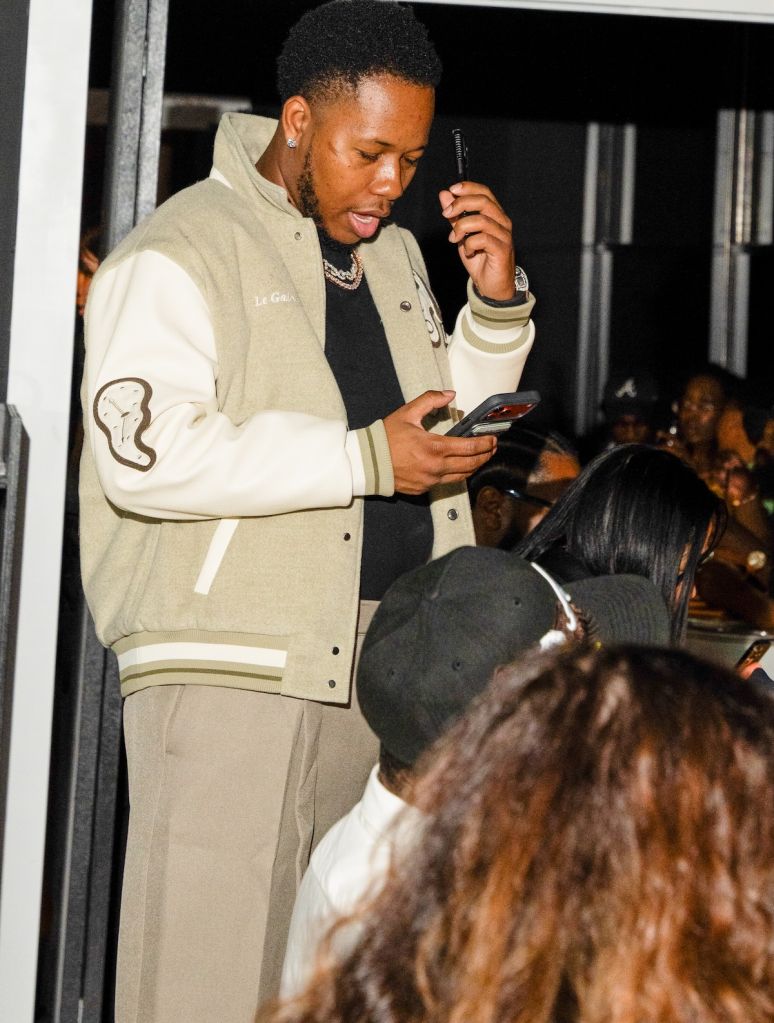 A person wearing a beige jacket and holding a smartphone, standing in front of a group of people.