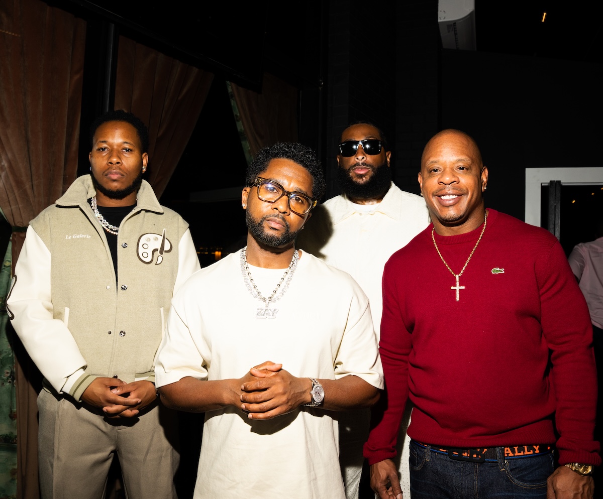 Four African American men standing together, wearing casual clothing and accessories including a cross necklace and sunglasses.