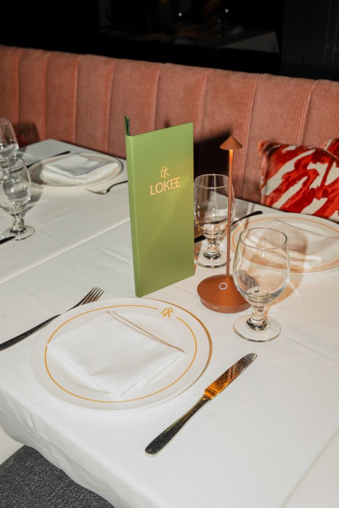A table setting at the Lokee restaurant, with a green menu, wine glasses, and a plate with utensils.