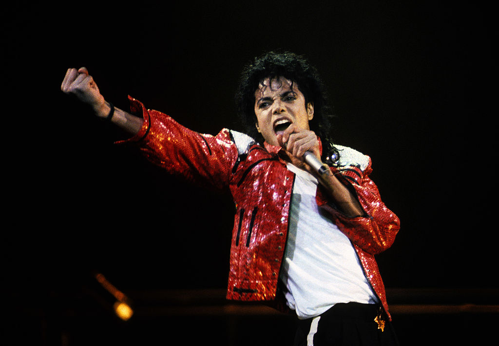 A performer in a red sequined jacket singing passionately on stage in a dark setting.