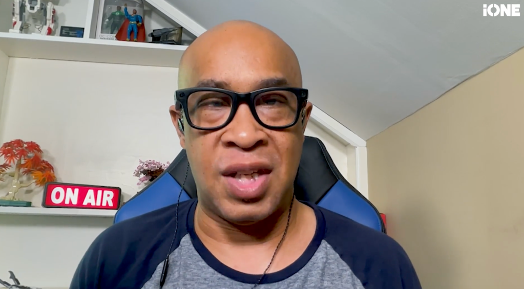 A bald man wearing glasses and a navy blue shirt, with an "ON AIR" sign visible behind him.