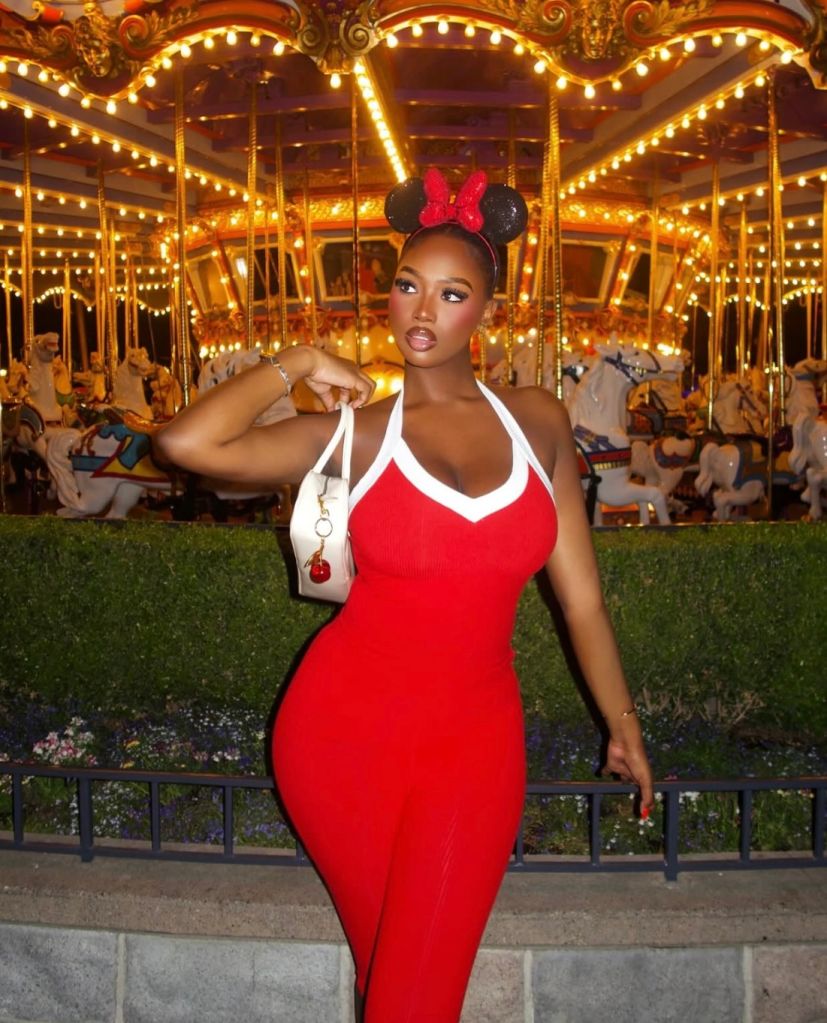 A woman in a red dress and Minnie Mouse-inspired headband posing in front of a colorful carousel at an amusement park.
