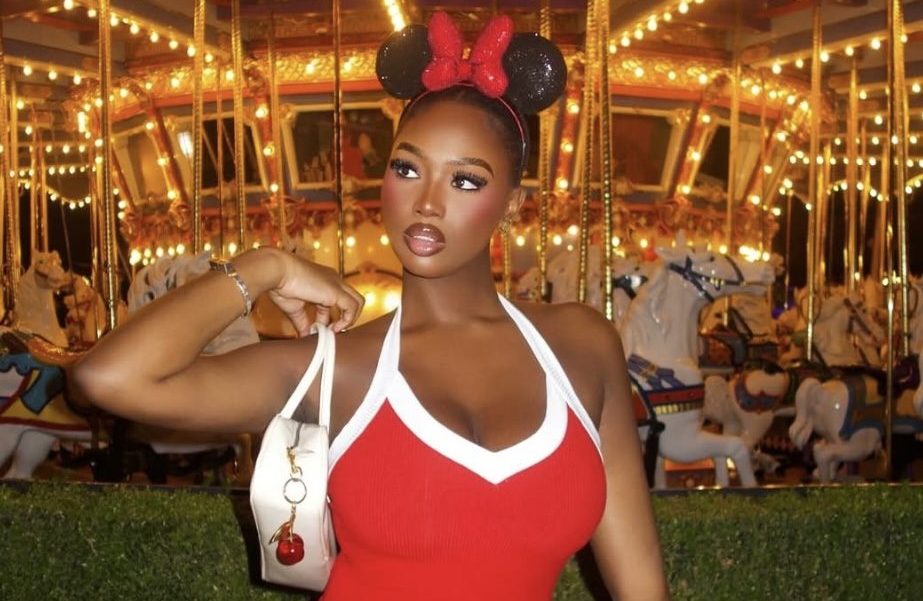 A woman wearing a red dress and Minnie Mouse-style ears stands in front of a brightly lit carousel in an amusement park setting.
