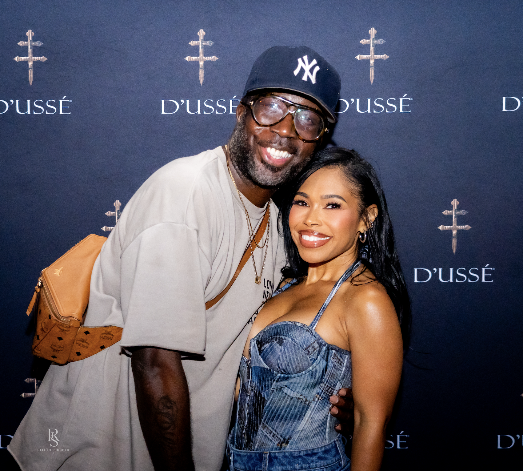 Two smiling people posing in front of a D'usse backdrop, with the man wearing a Yankees baseball cap and the woman wearing a denim jumpsuit.