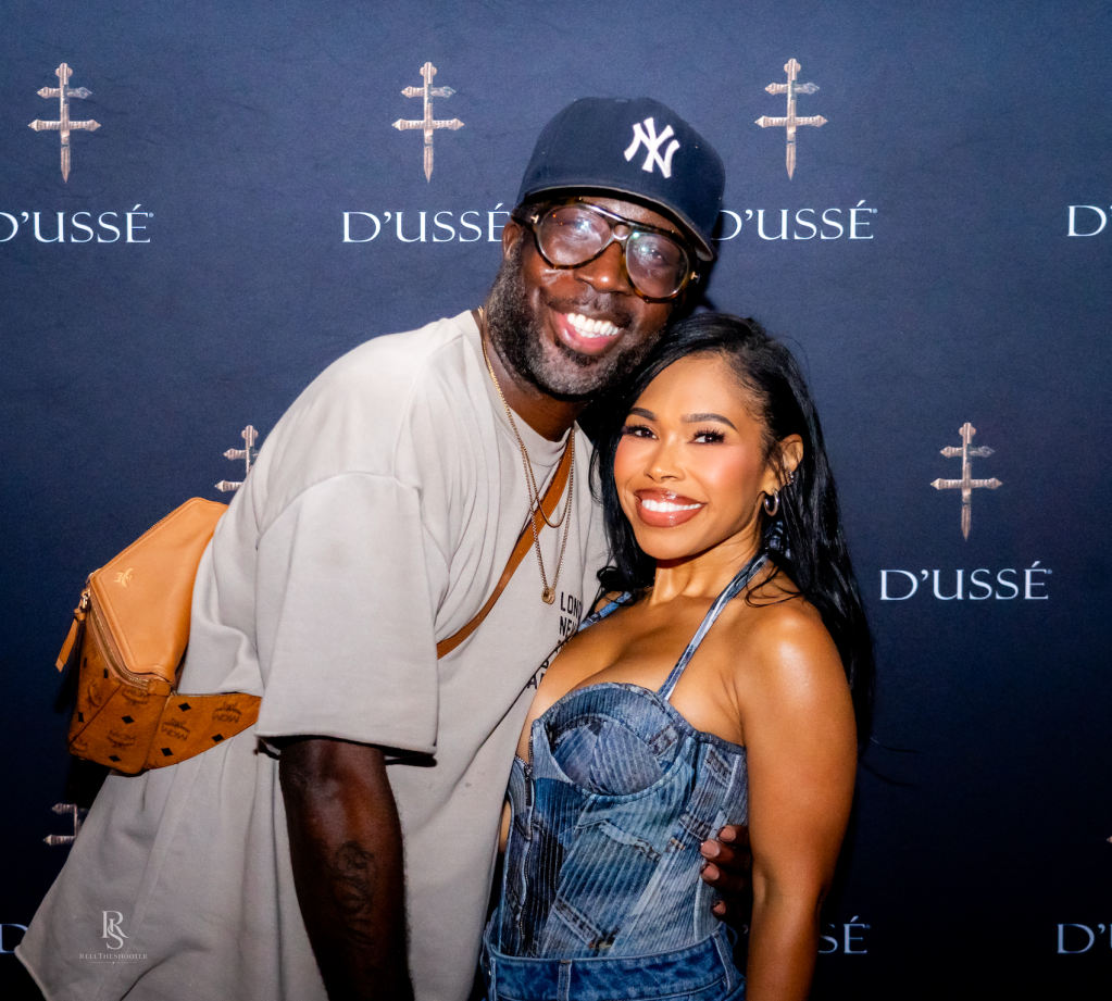 Two smiling people posing in front of a D'usse backdrop, with the man wearing a Yankees baseball cap and the woman wearing a denim jumpsuit.