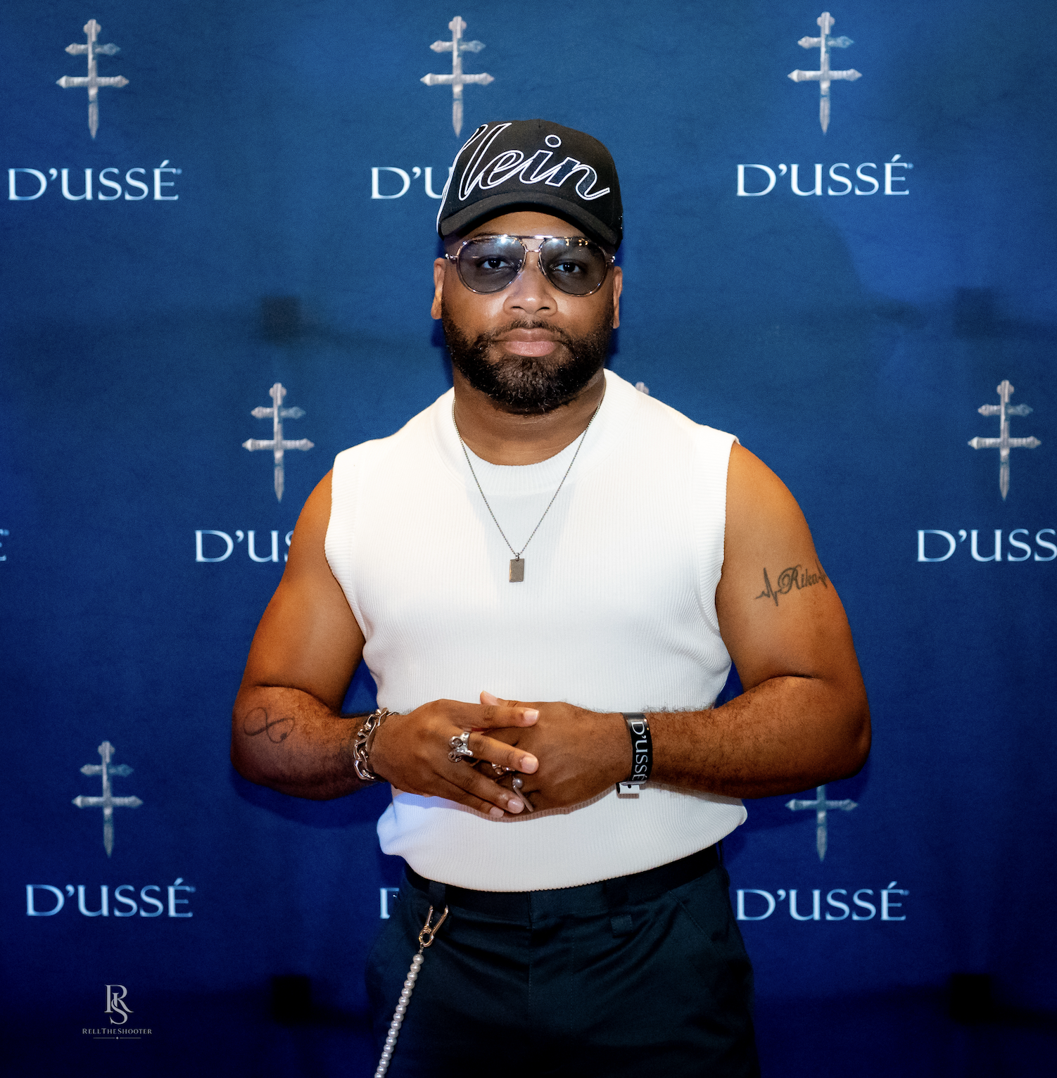 A man wearing a black hat with "D'USSE" text and sunglasses, standing in front of a blue backdrop with D'USSE branding.