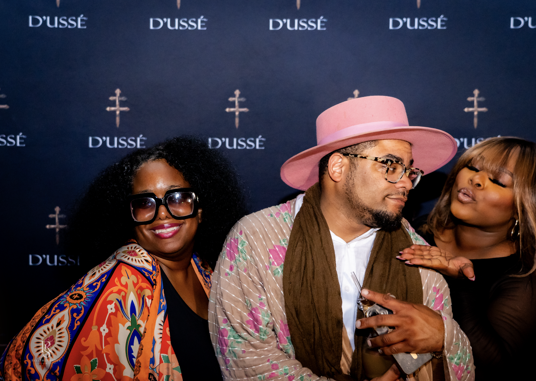 Three people posing in front of a D'usse backdrop, one wearing a pink hat and sunglasses, another wearing a patterned shawl, and the third with a beard and glasses.