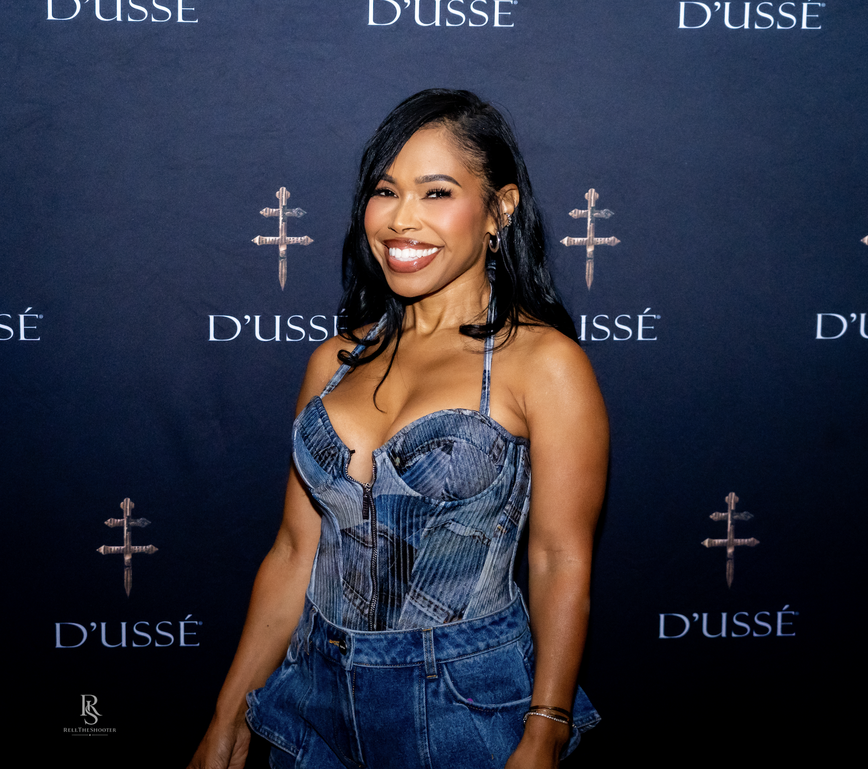 A smiling woman wearing a denim jumpsuit poses in front of a backdrop with the "D'usse" logo.
