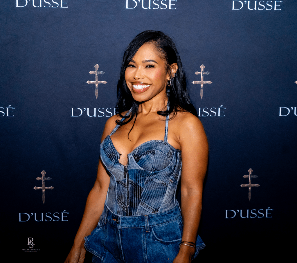 A smiling woman wearing a denim jumpsuit poses in front of a backdrop with the "D'usse" logo.